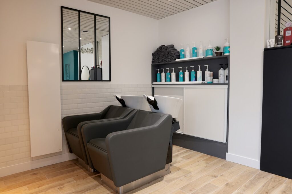 Bacs de lavage gris et étagères de soins capillaires dans un salon de coiffure moderne au sol boisé.