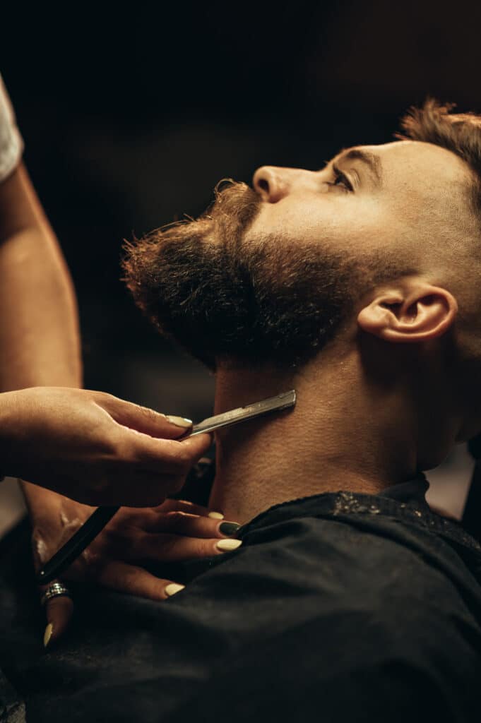 Barbier rasant de près le cou d'un homme barbu avec un rasoir coupe-chou sous une lumière chaude.